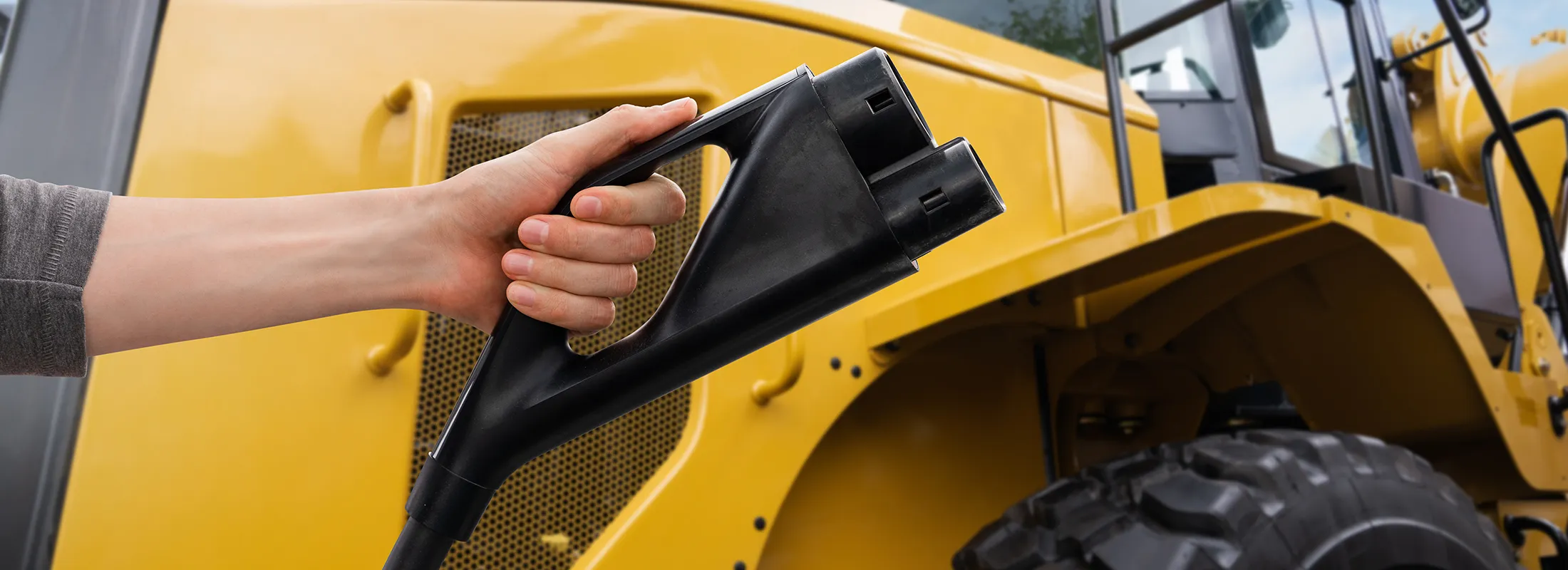 Close-up of a hand holding a large black electrical connector, with a tractor in the background
