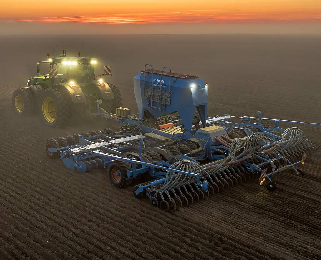 Tractor pulling a large blue seed drill across a field at sunset.